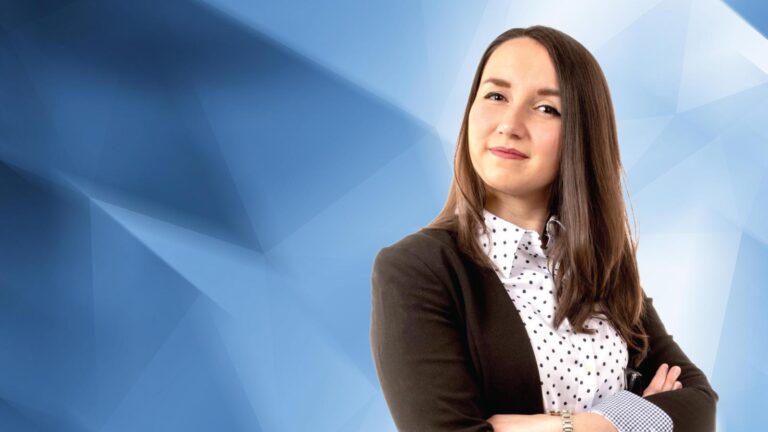 A woman with long brown hair is standing with her arms crossed, wearing a white polka-dot shirt and a dark blazer, against a blue geometric background.