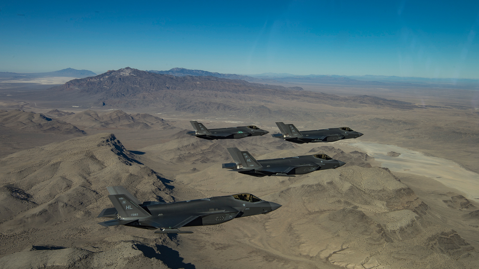Four military fighter jets in formation fly over a mountainous desert landscape under a clear blue sky.