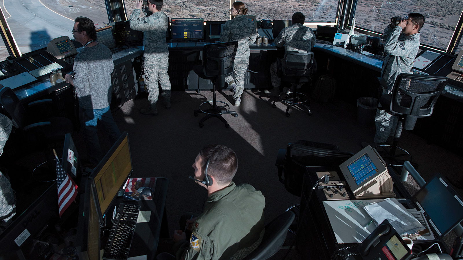 Military personnel and civilians work inside an air traffic control tower, monitoring screens and using binoculars to oversee runway operations.