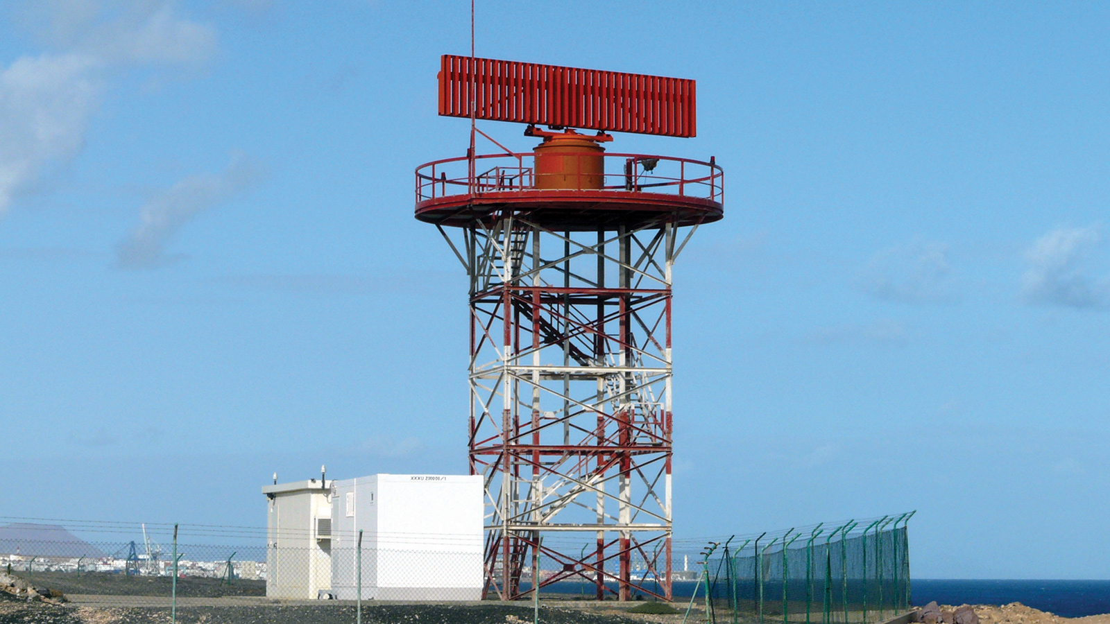 A tall, red-and-white radar tower stands on a concrete base with a small white building nearby, surrounded by a fence. The radar is set against a backdrop of clear blue sky.