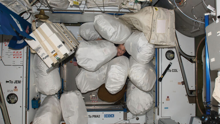 An individual smiling and partially obscured by several large white storage bags inside what appears to be a space station module.
