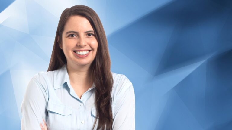 A woman with long brown hair, wearing a light blue shirt, smiles and stands with her arms crossed against a blue geometric background.