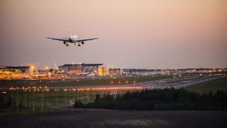 An airplane prepares to land on a runway at an airport during twilight, with airport buildings and lights visible in the background.