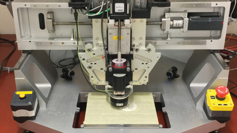 Close-up of a 3D printer with intricate components, showing its print head in the center, wiring, and control buttons on the sides. The machine is located on a solid workbench surface.
