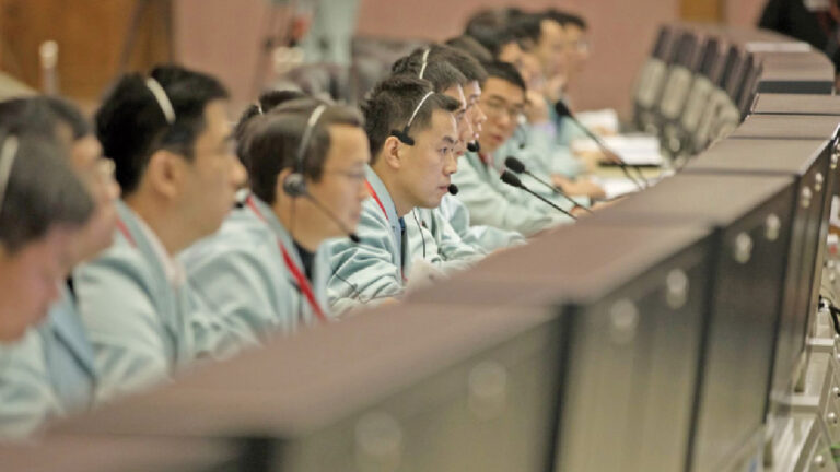 A group of people wearing headsets and blue jackets sit at a row of computer monitors, attentively working or listening.