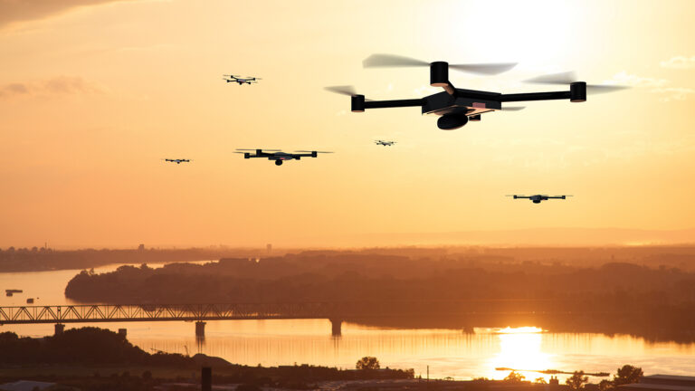 Several drones fly over a river and bridge at sunset, with an orange sky and distant landscape in the background.