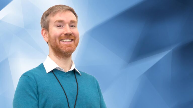 A man with a beard and a smile stands against a blue geometric background. He wears a teal sweater over a white collared shirt and has a black lanyard around his neck.