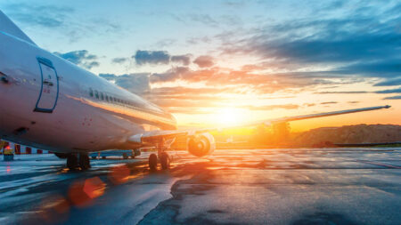 A commercial airplane is parked on a wet tarmac at sunrise, with the sun shining through scattered clouds in the background.