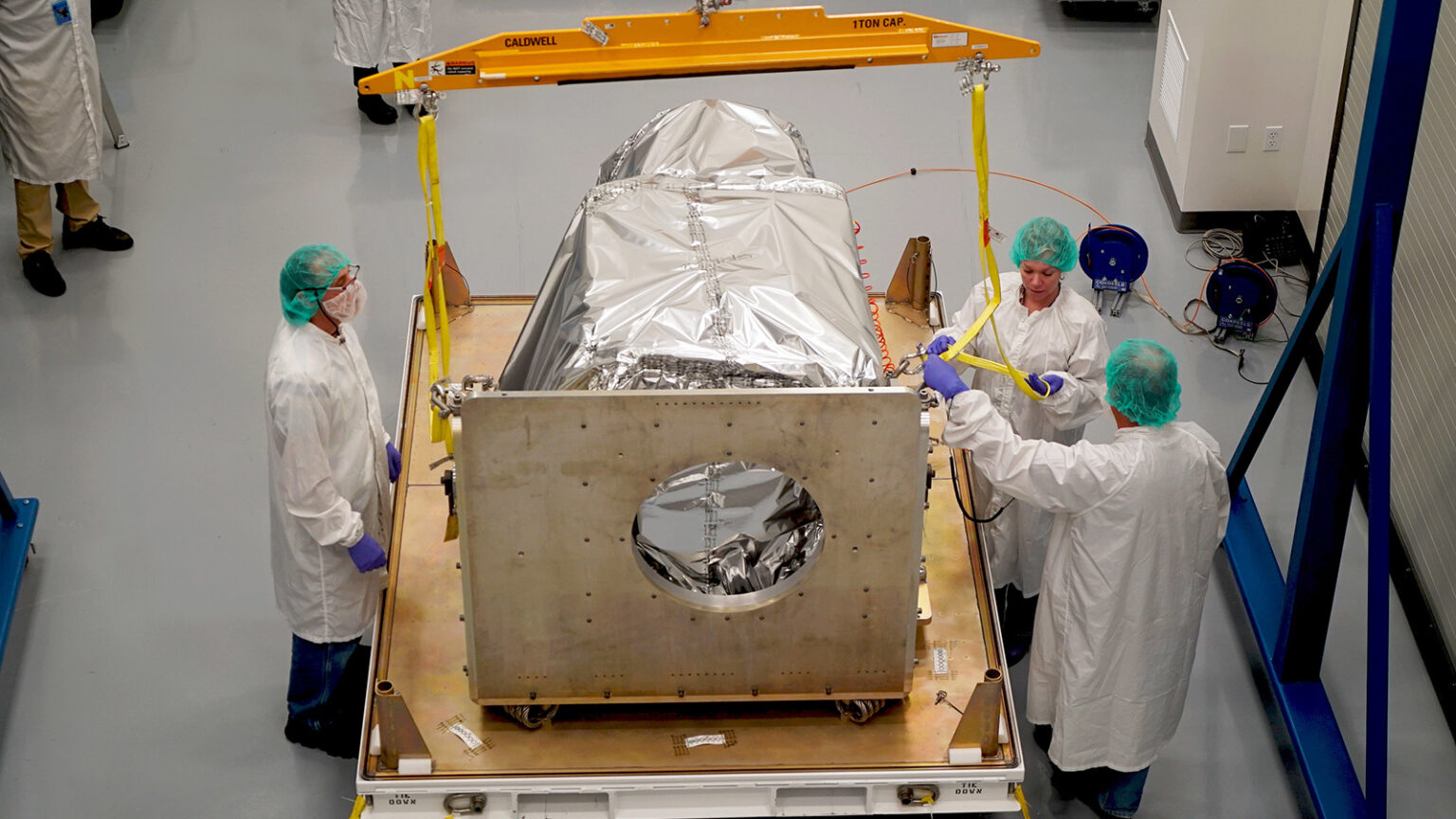Three technicians in lab coats, gloves, and hairnets work on securing a silver foil-wrapped object suspended by a yellow gantry in a cleanroom.