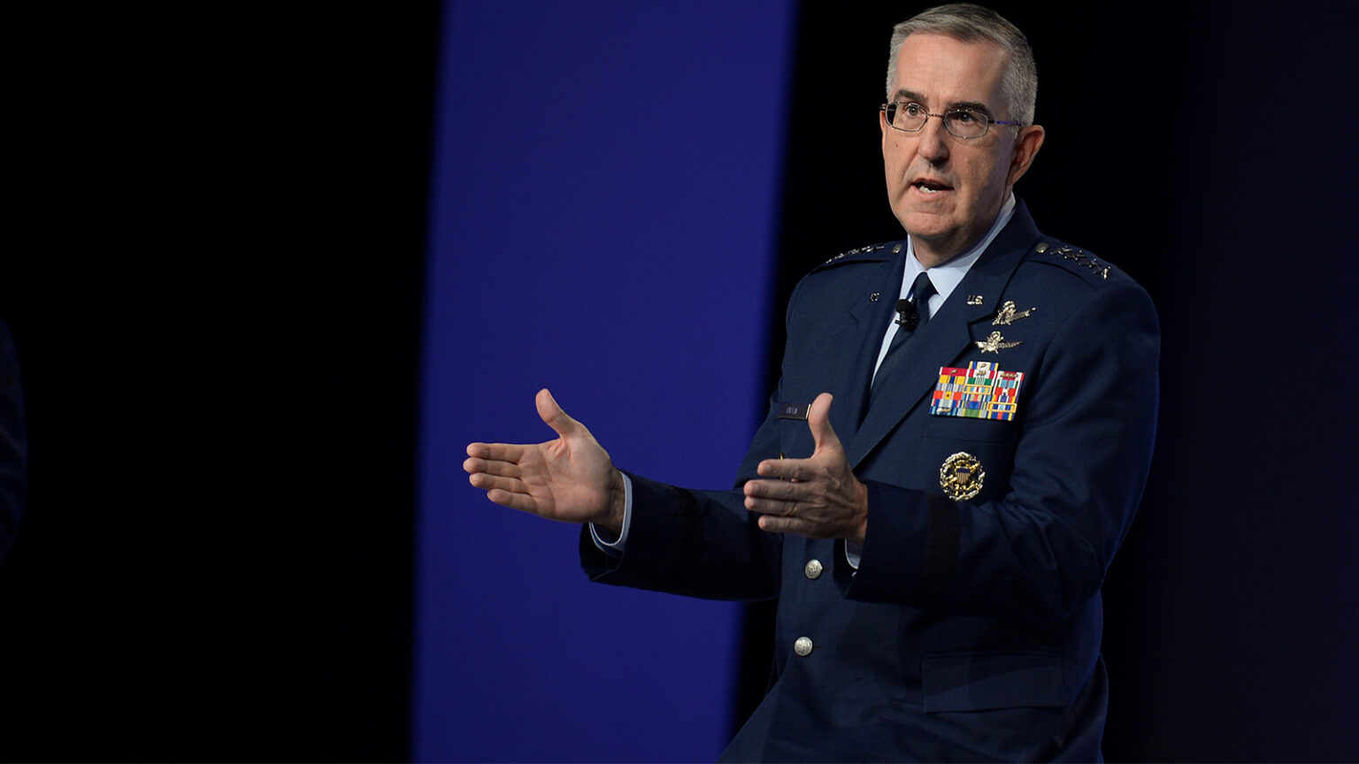 A person in military uniform with numerous medals and insignia gestures with both hands while speaking against a blue backdrop.