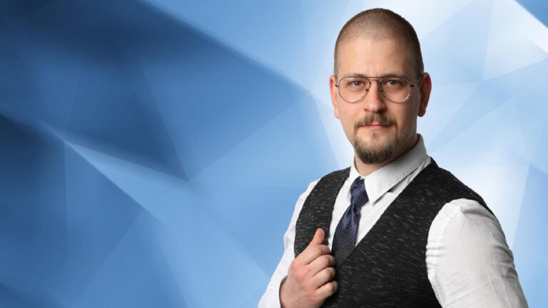 A person with glasses, a beard, and short hair, wearing a white shirt, tie, and dark vest, stands against a geometric blue background.