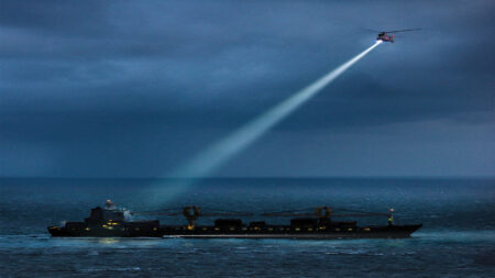 A helicopter shines a spotlight on a large cargo ship in the ocean under a cloudy, dusk sky.