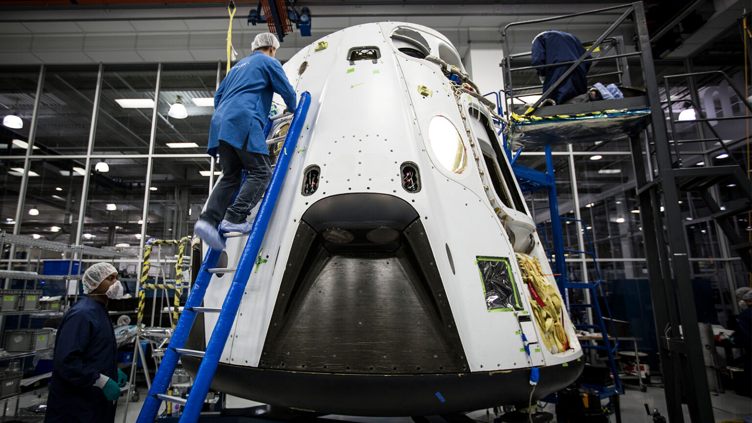 Technicians work on a space capsule inside a high-tech facility, standing on ladders and wearing protective gear. The capsule is partially assembled and surrounded by various tools and equipment.