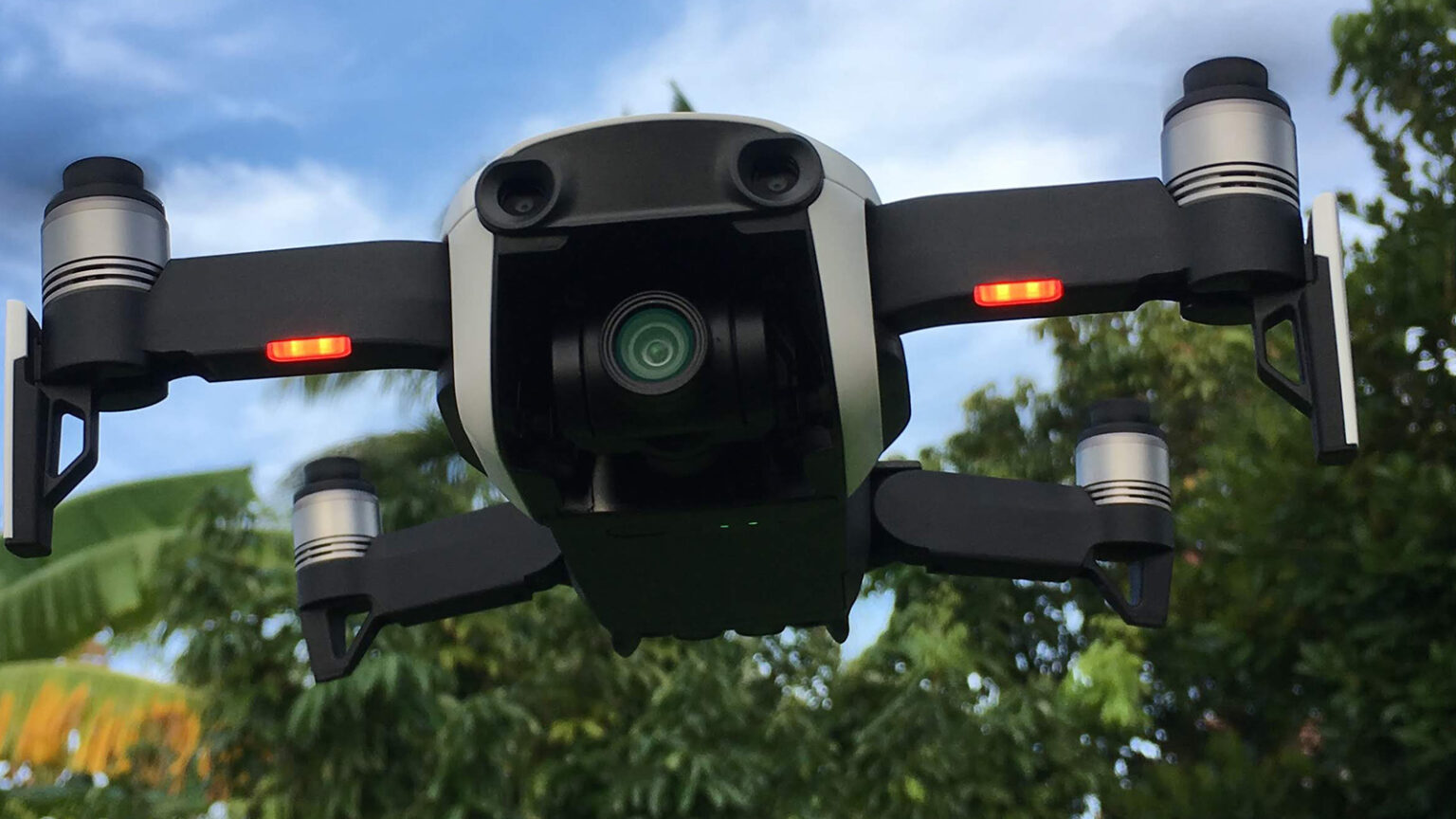 A drone with a camera hovers in mid-air against a background of trees and a cloudy sky.