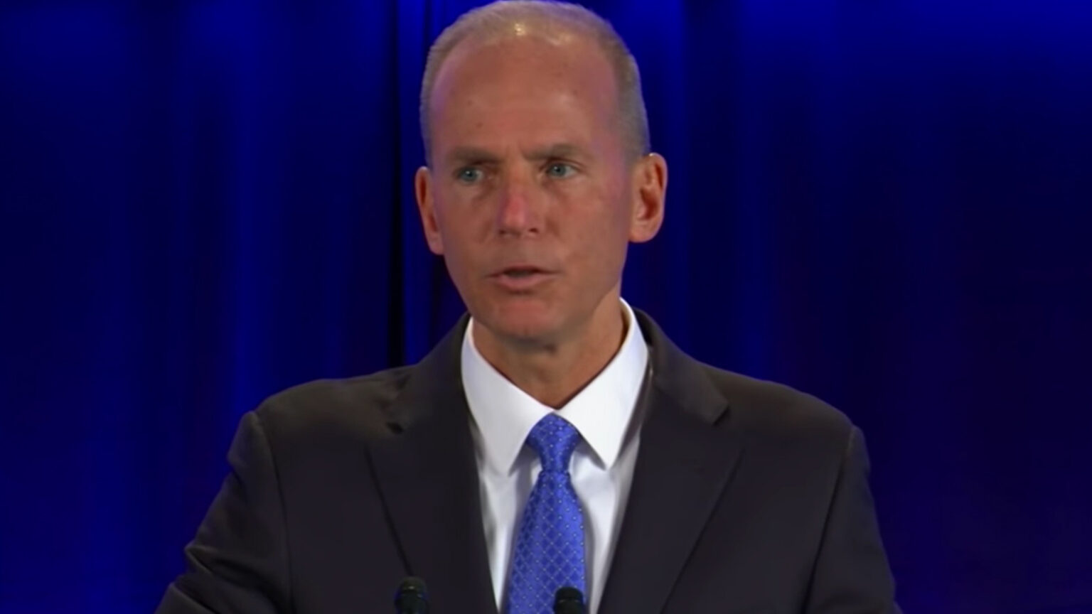 A man in a suit and blue tie is speaking at a podium with a blue curtain in the background.