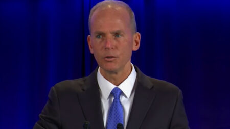 A man in a suit and blue tie is speaking at a podium with a blue curtain in the background.