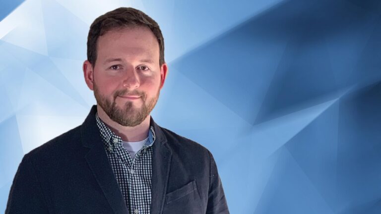 A man with a beard and mustache, wearing a black blazer over a checkered shirt, stands against a background with shades of blue.