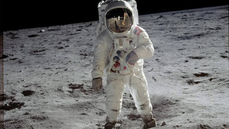 An astronaut in a spacesuit stands on the moon's surface with footprints visible in the dusty ground.