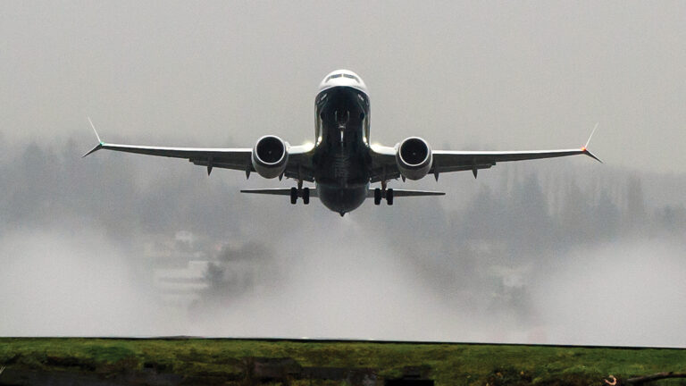 A commercial airplane ascends into the sky from a runway, generating visible water vapor or exhaust trails in the misty backdrop.