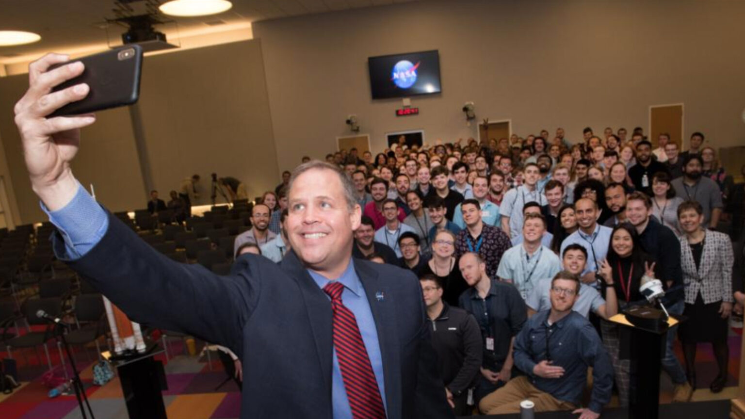 A man is taking a selfie with a large group of smiling people in a conference room. A NASA logo is visible on a screen in the background.