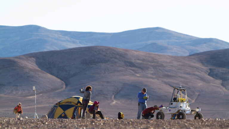 Several people in a desert setting work around a yellow tent and a small rover-like vehicle with hills in the background.