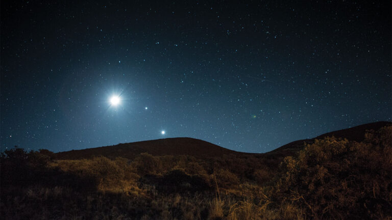 A night sky filled with stars over a hilly landscape, with a particularly bright celestial object near the horizon.