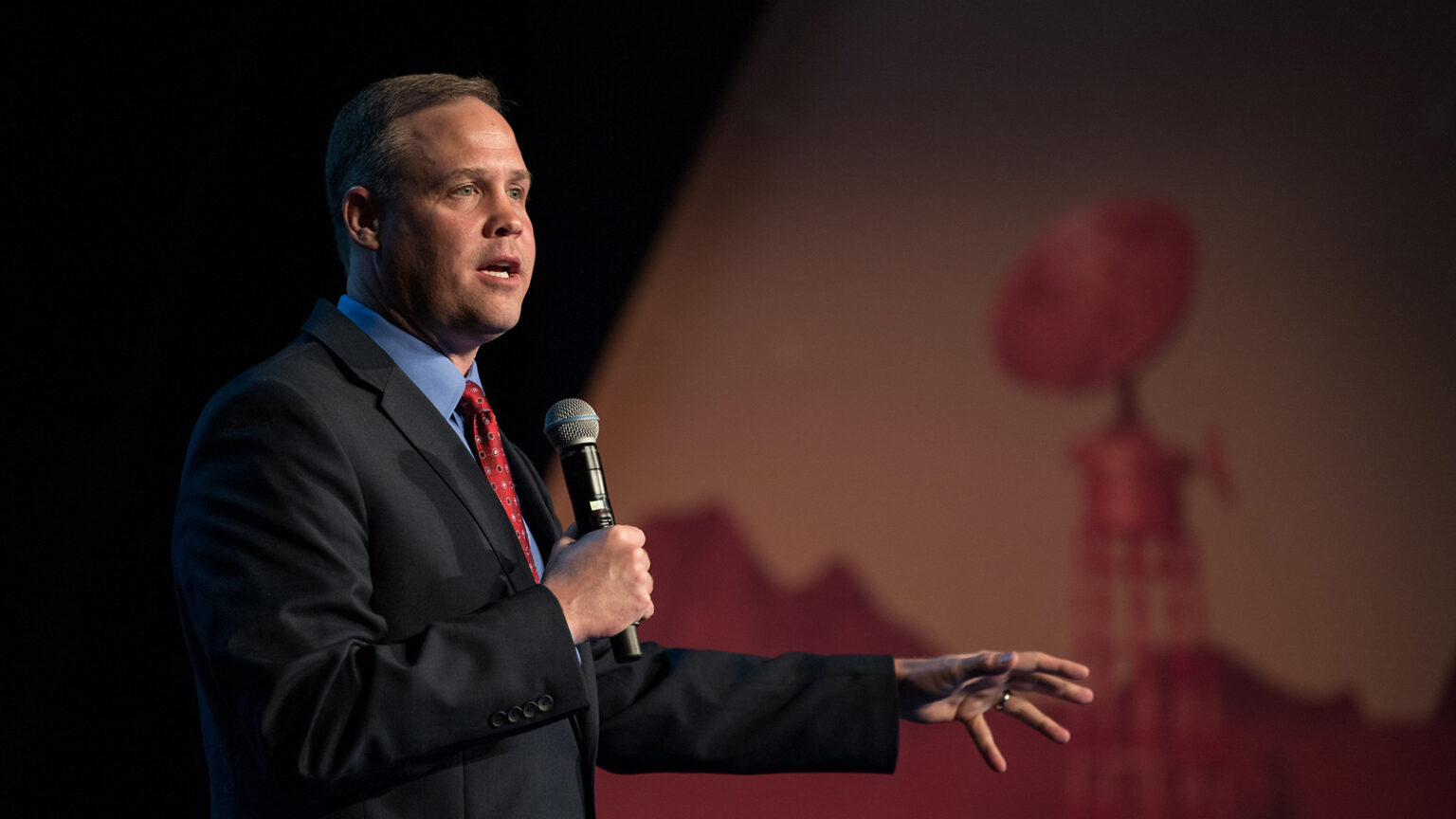 A man in a suit and tie speaks into a microphone on a stage, gesturing with one hand. A background image features a communication dish and mountain silhouettes.