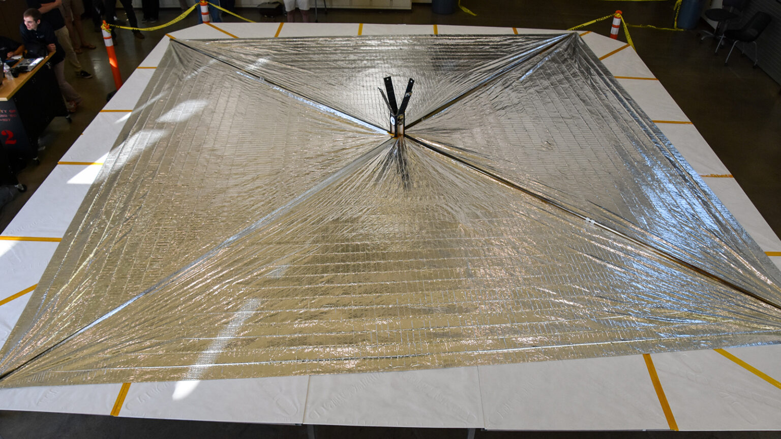 A large, reflective solar sail is fully deployed and displayed indoors, surrounded by caution tape and cones. People are observing from the left.