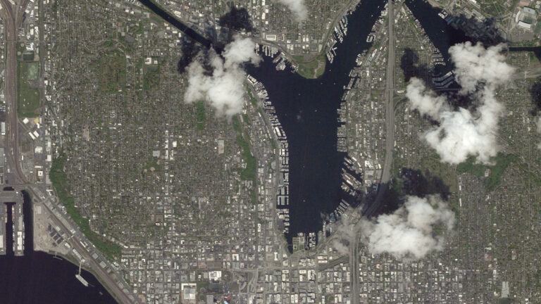 Aerial view of a city featuring a narrow waterway running vertically down the center, surrounded by urban buildings and green areas, with scattered clouds partially obstructing the view.