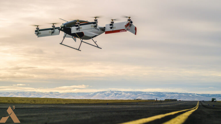 A large drone with multiple propellers flies over a runway in a vast, open landscape with mountains in the background.