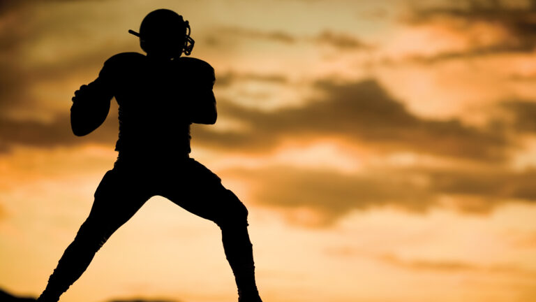 Silhouette of an American football player standing against a sunset sky, wearing a helmet and uniform.