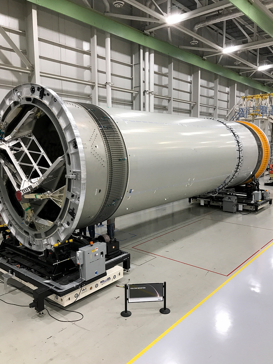 A large rocket component lies horizontally on a wheeled platform inside a spacious industrial facility, surrounded by machinery and equipment.