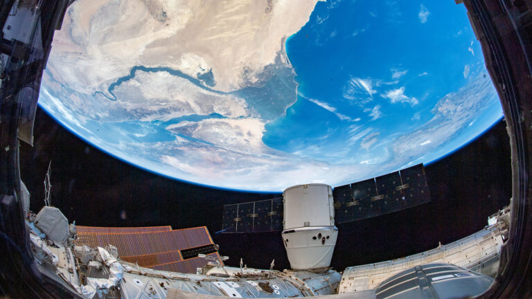 Image taken from the International Space Station showing the Earth’s curved horizon, with a module and solar panels in the foreground and a coastline below.