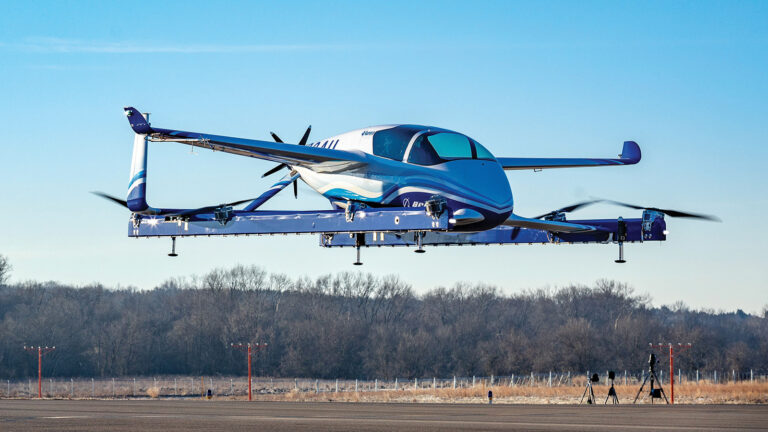 A sleek, futuristic electric aircraft hovers mid-air over a runway with trees in the background on a clear day.