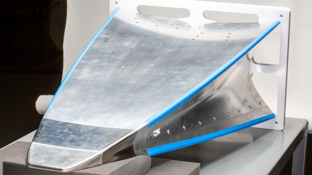 A metal aerofoil section with blue tape along the edges is mounted on a white frame, positioned on a gray cushioned surface in a well-lit indoor setting.
