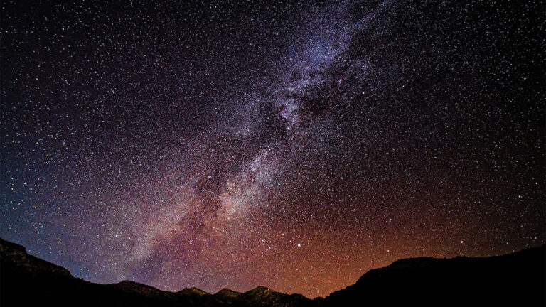 A clear night sky showcases the Milky Way galaxy above a silhouetted mountain range, with countless stars illuminating the scene.