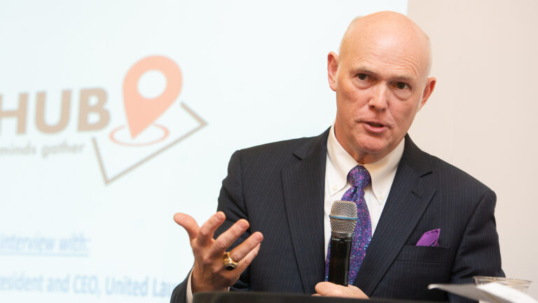 A bald man in a suit and purple tie speaks into a microphone while gesturing with his hand. A presentation screen with partial text "HUB" is visible behind him.