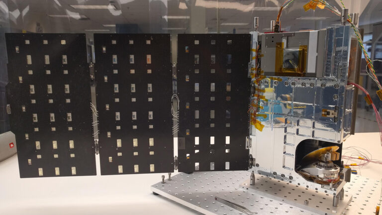 A small satellite prototype with extended solar panels is displayed on a table. The main structure is metallic with visible wiring and components.