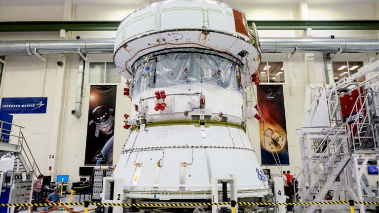 A spacecraft module under construction in an industrial facility with engineers working nearby. Various equipment and machinery are visible around the module.