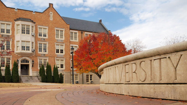 A university building with a classic brick facade is surrounded by fall foliage. A stone wall with the word "UNIVERSITY" etched into it is visible in the foreground.