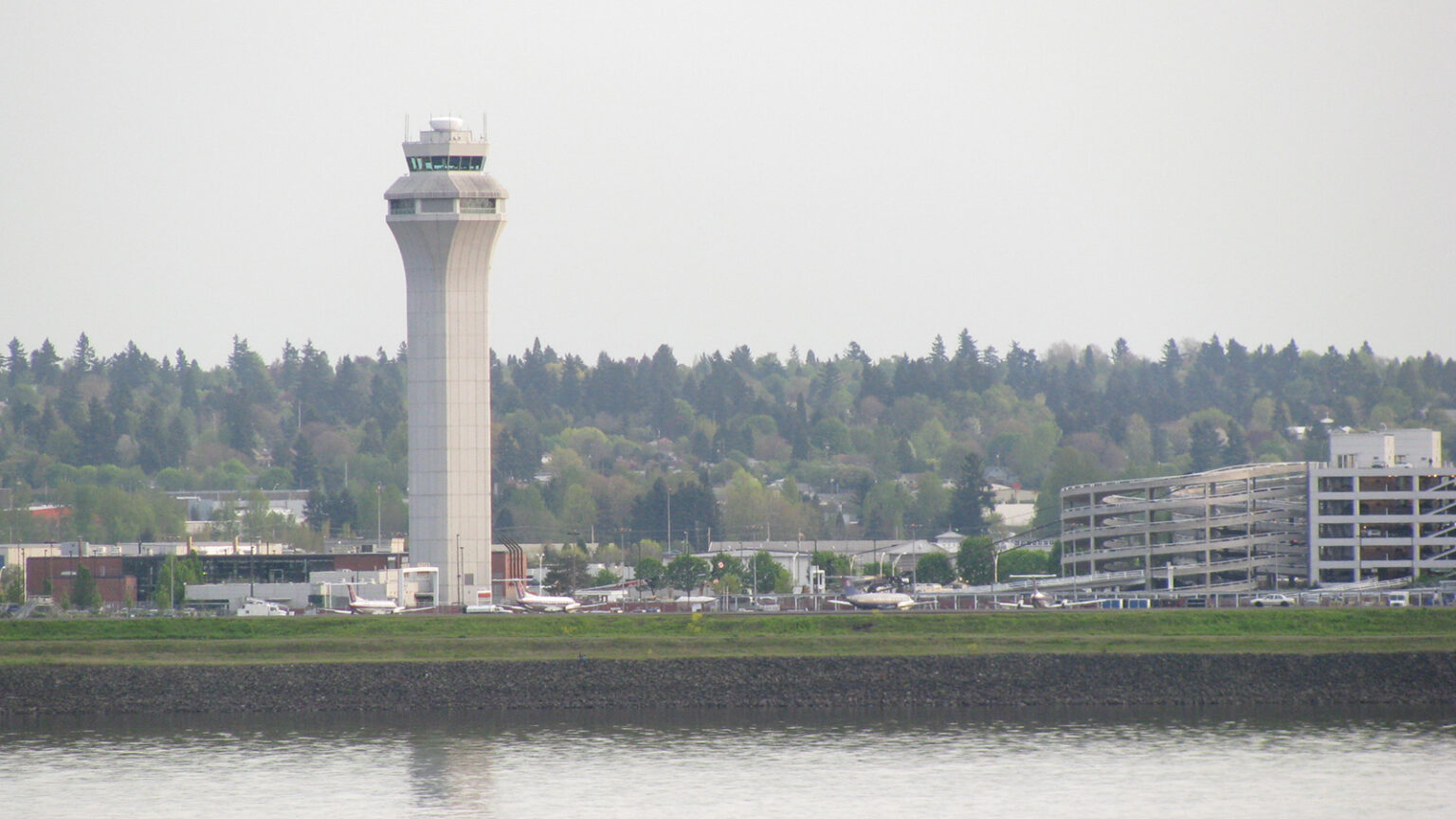 Image of an airport control tower next to a parking structure with a river in the foreground and a forested area in the background.