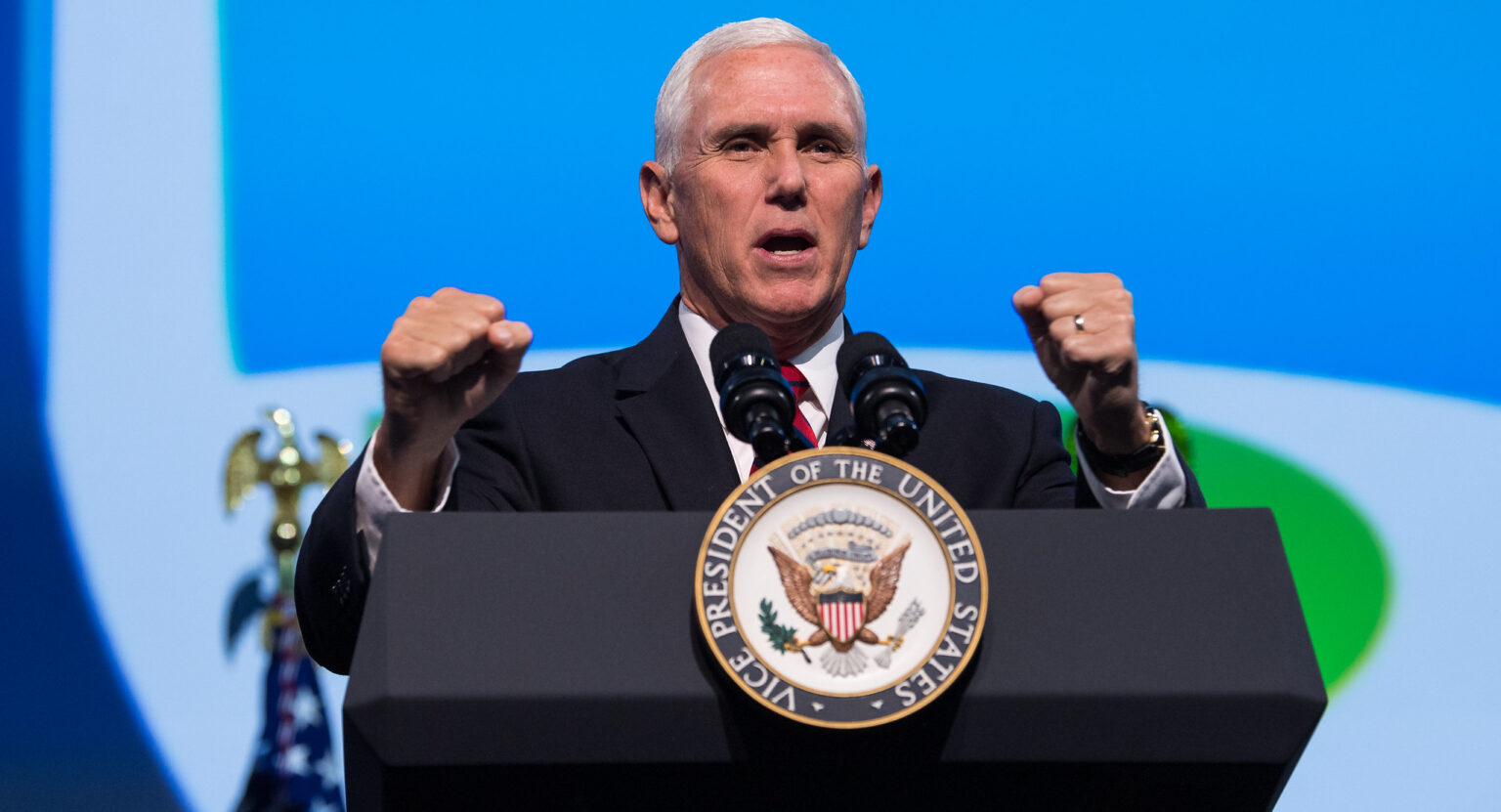 A man in a suit and tie stands at a podium with the emblem of the Vice President of the United States. He has his fists raised slightly and appears to be speaking. The background features a blue and green design.
