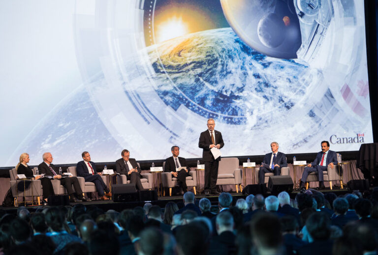 People in formal attire sit on a stage discussing space exploration, with an image of Earth and a spacecraft projected in the background. A speaker stands at a podium. The audience faces the stage.