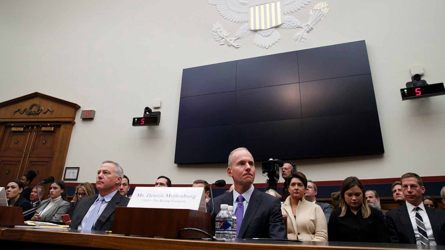 A group of people, some seated at desks, attend a session in a formal room with a large screen and the U.S. Seal on the wall. A nameplate for “Mr. Dennis Muilenburg, CEO – The Boeing Company” is visible.