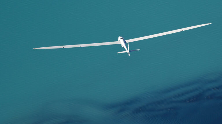 A white glider with long wings is flying over a body of turquoise water, casting a shadow below.