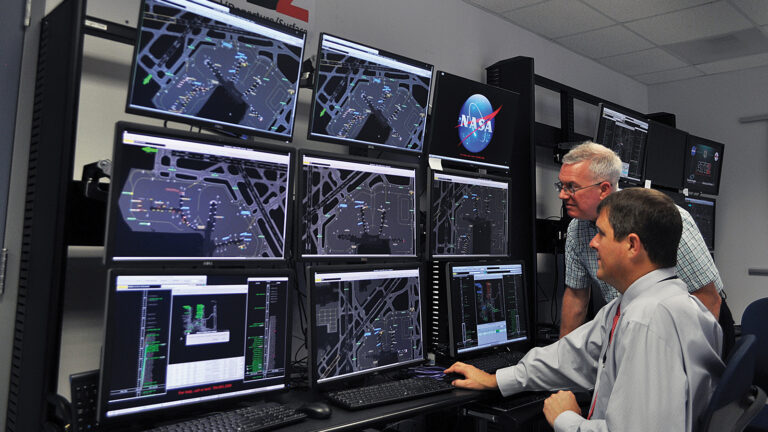 Two men monitor multiple screens displaying airport layouts and data in a NASA control room.
