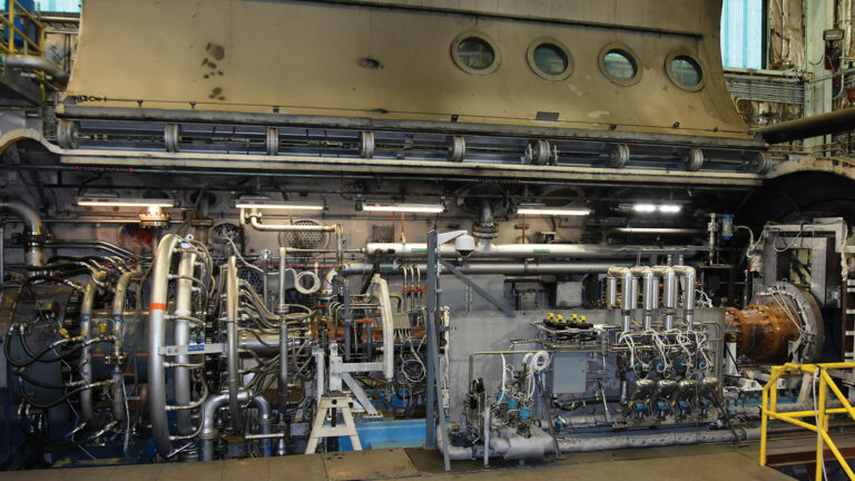 Close-up view of industrial machinery with numerous pipes, valves, and components against a large metal structure in an engineering or manufacturing facility.