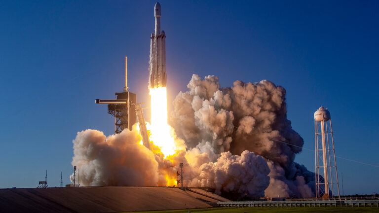 A rocket lifting off from a launchpad with flames and smoke billowing out from the base against a clear blue sky backdrop.