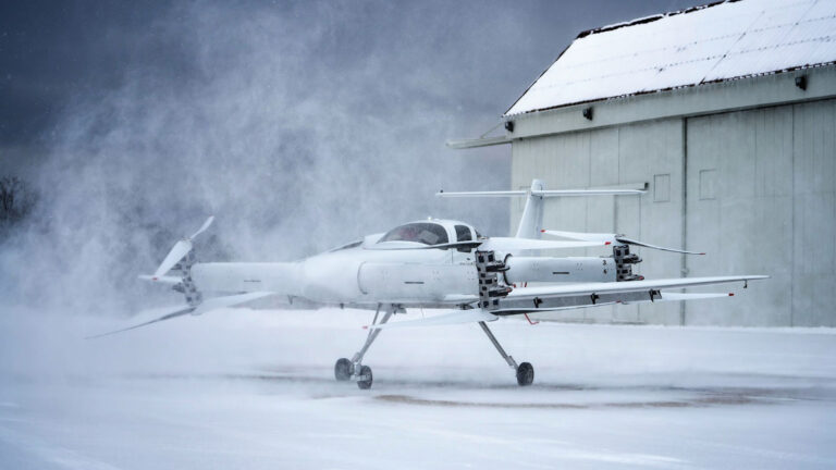 A white unmanned aerial vehicle with propellers on its wings rests on a snow-covered ground in front of a large hangar under a cloudy sky, with snow being blown around.