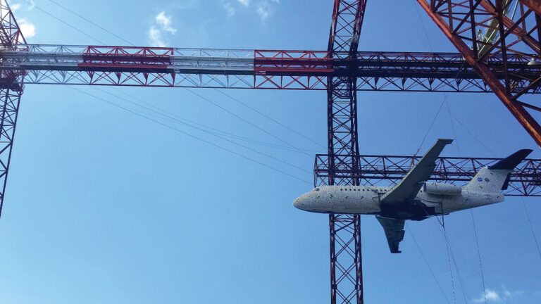 A large airplane is suspended from red and white gantries against a clear blue sky, appearing to be part of a structural or artistic installation.